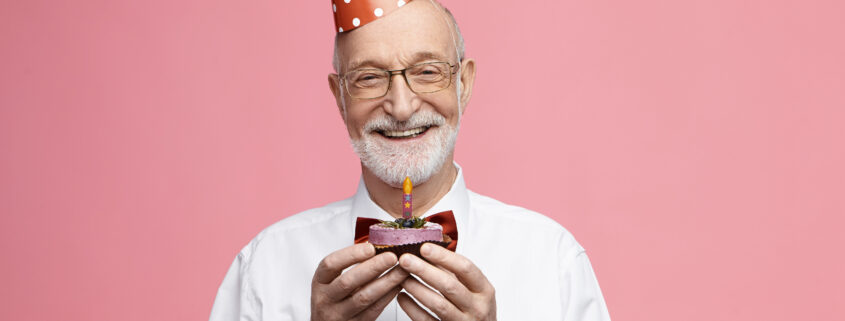 Attractive happy retired Caucasian male wearing bow tie, glasses and cone hat celebrating his 80th anniversary, posing isolated with birthday cake in his hands, going to blow out candle and make wish