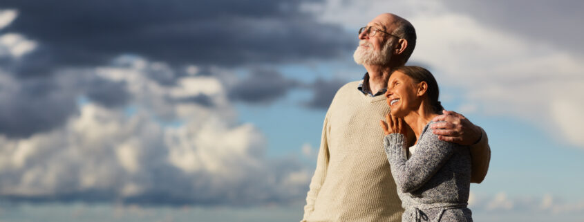 Amorous seniors standing by water against stormy sky and enjoying sundown
